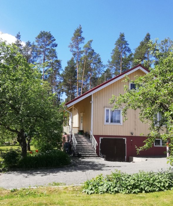 Gult trähus med rött garage, omgivet av grönska och träd under en klarblå himmel.