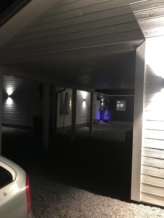 Mörk vy av en carport upplyst av flera väggmonterade spotlights, med en julgran synlig i bakgrunden.