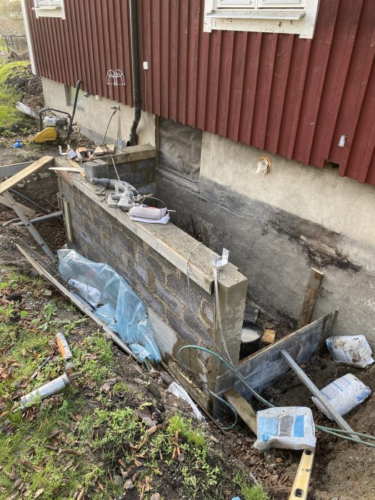 Byggplats med nyligen gjuten betongplatta och mur vid en röd husvägg, verktyg och material utspridda på marken.