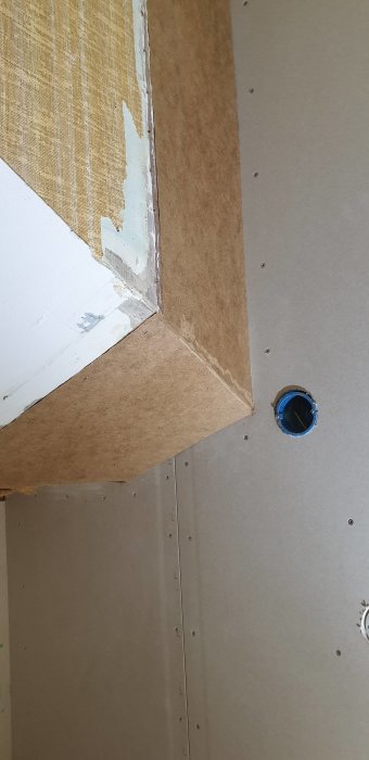 Renoverad vägg med ny masonit och delvis borttagen tapet längs ett hörn, ovanför en elektrisk uttag.