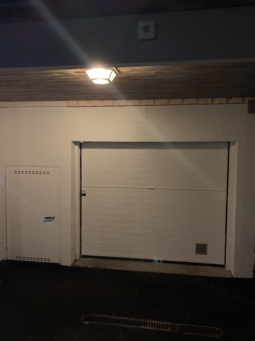 Garageport upplyst av en takspotlight på en vitmålad betongvägg under natten.