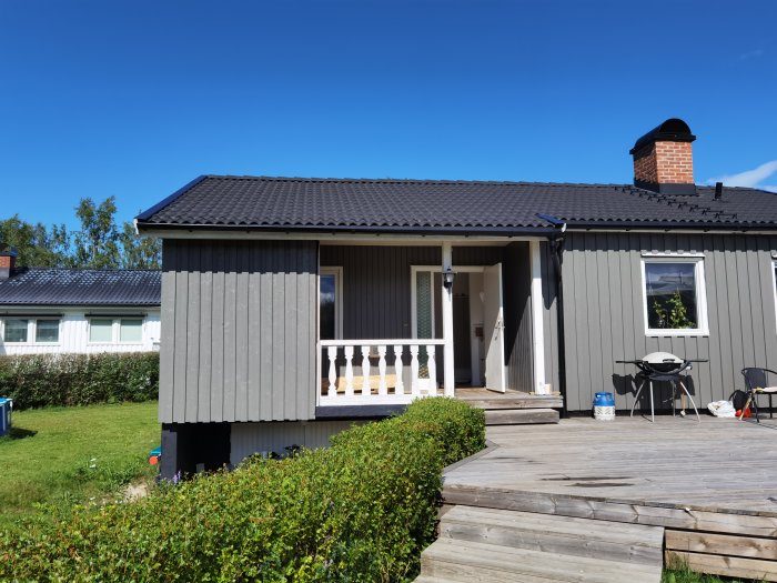 Ett enfamiljshus med mörkgrå panel och svart tak, en veranda med vita staketstolpar och en trätrall framför.