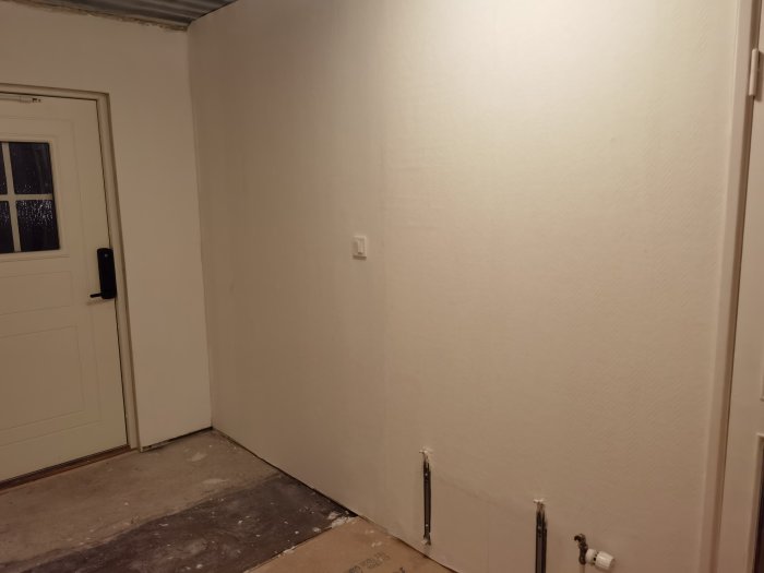 Hörn av ett rum under renovering med vit vägg, kartong på golvet och avlägsnade element.
