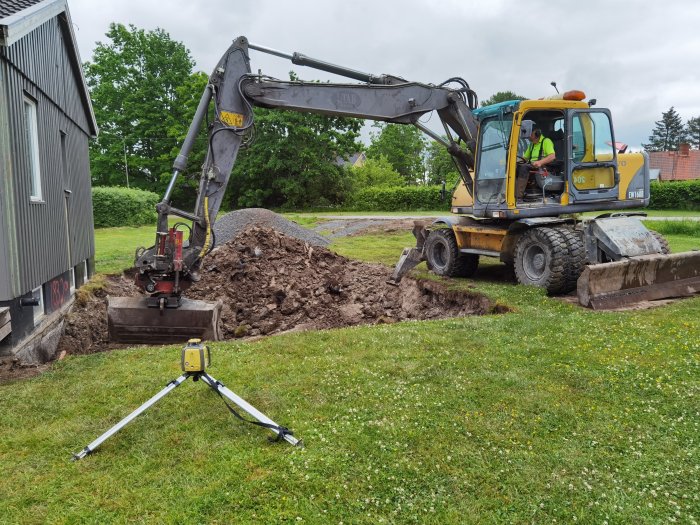 Grävmaskin gräver ut jord nära en byggnad med en hög grushög och en nivelleringslaser på stativ i förgrunden.
