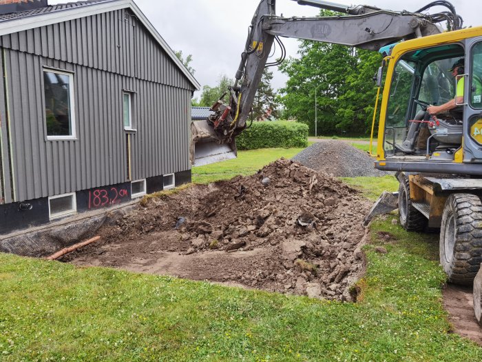 Grävmaskin som gräver upp jord bredvid hus med högen grus i bakgrunden.