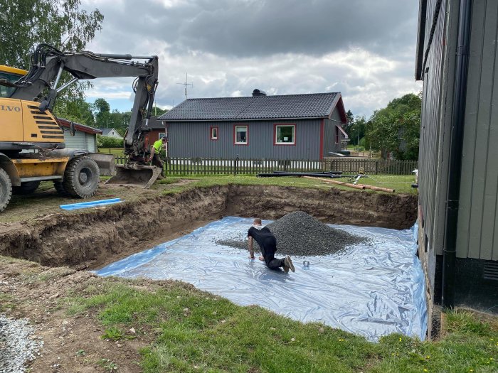 Grundarbete för husbygge med grävmaskin, hög med grus och arbetare som förbereder marken.