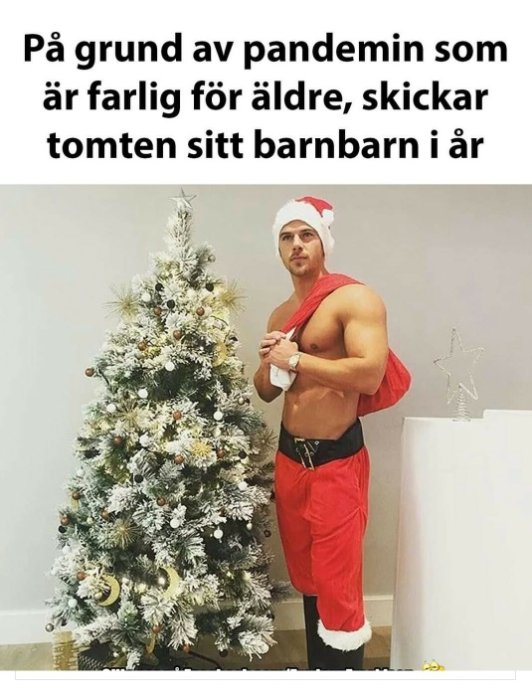 Man i tomtedräkt och tomteluva poserar med muskler bredvid en julgran.