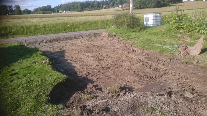 Nygrävd infart till en gårdsplan med synlig grushög och en uppgrävd stubbe vid en grävmaskinsskopa, omgivet av gräsmark.