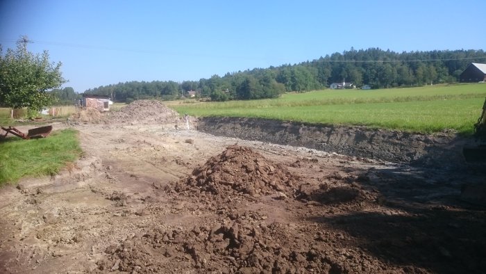 Nygrävd infart i en gårdsplan med jordhögar och en grävmaskin, omgiven av gröna fält och en ladugård i bakgrunden.