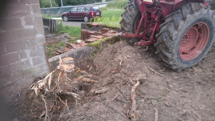 Traktor bredvid en utgrävd stubbe vid en byggnad, med uppgrävd jord och tegelstenar runtomkring.