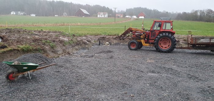 Bild av en röd traktor med kärra bredvid nygrävd infart täckt med grus, på en gård med utsikt över åkrar.