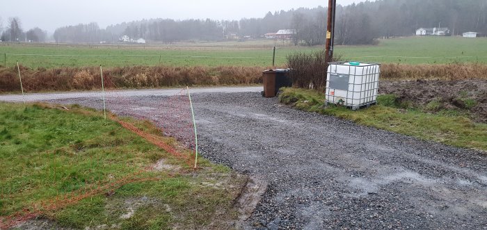 Nygrävd infart till gårdsplan med grus och markeringsstolpar, i lantlig miljö under regnigt väder.