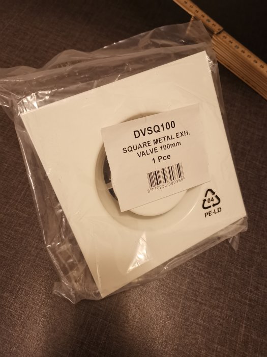 Förpackat fyrkantigt frånluftsdon märkt "DVSQ100 SQUARE METAL EXH. VALVE 100mm" bredvid ett måttband.