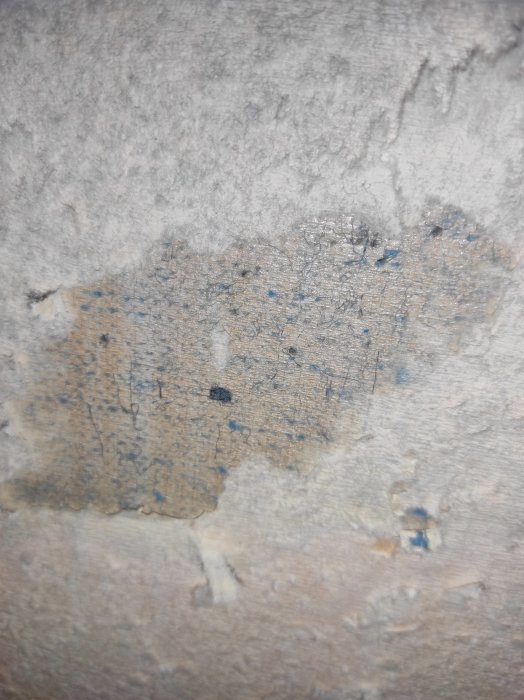 Avskalade tapetrester på en vägg med synlig struktur, eventuellt gammalt klister eller underlager.