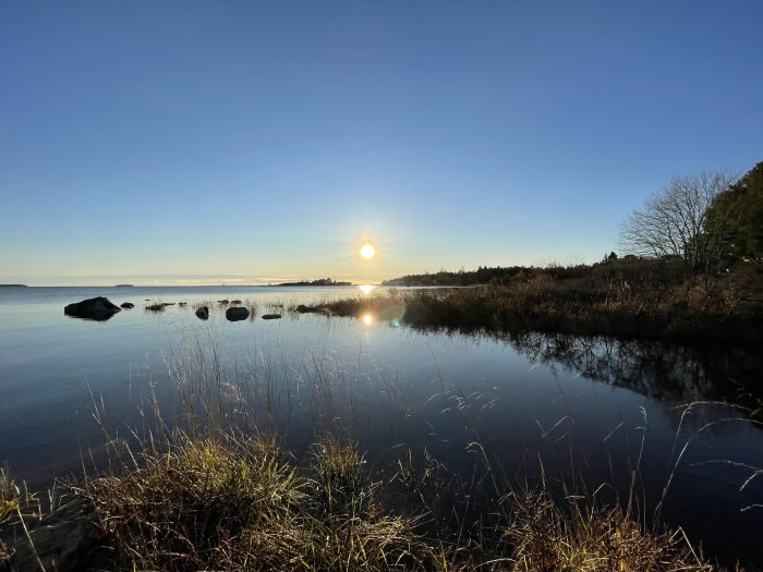 Solnedgång vid en stilla sjö med stenar och växtlighet, spegling av solen i vattnet.