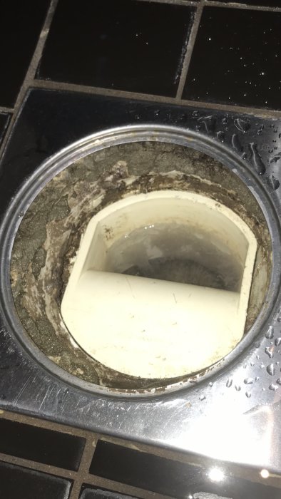 Golvbrunn i badrum utan synlig manschettring, omgiven av mycket fix och mörka plattor.