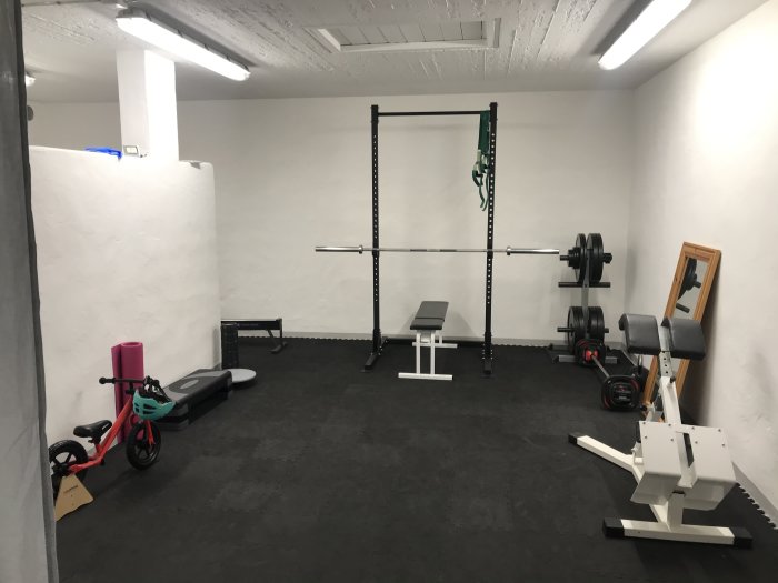 Målat gymrum med grova vita väggar, skivstångsställning & träningsutrustning, matt tak och belysning.