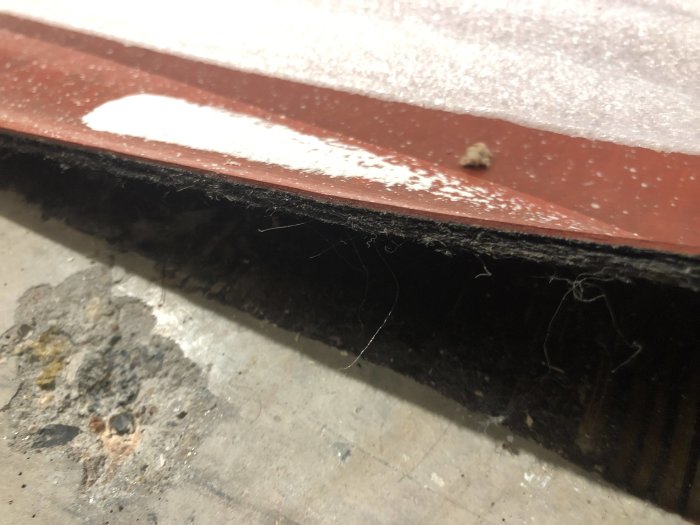 Gammal plastmatta delvis avtäckt under revet parkettgolv, ligger på betonggrund med synliga dammpartiklar och smuts.