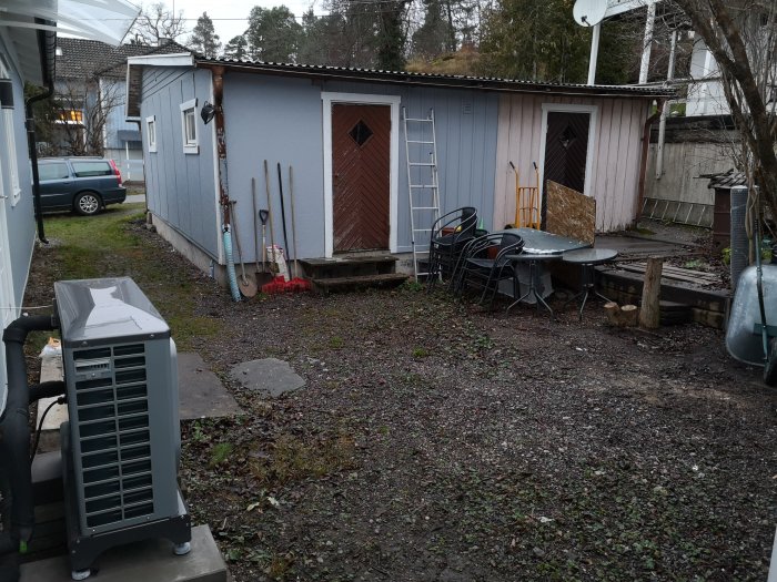 Trött uteplats med grus, trädgårdsmöbler och en delvis synlig värmepump, mellan ett blått hus och en röd bod.