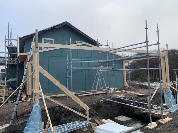 Byggplats med påbörjad carportkonstruktion vid isolerad gjutning och hus med ställningar.