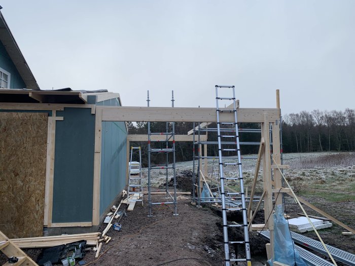 Pågående bygge av carport med isolerade stolpar och delvis uppsatt takstruktur, omgiven av stegar och byggmaterial.