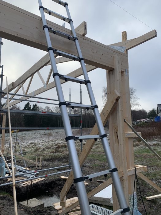 Trästolpar och en ställning i förgrunden med påbörjat tak av träkonstruktion till en carport under uppbyggnad.