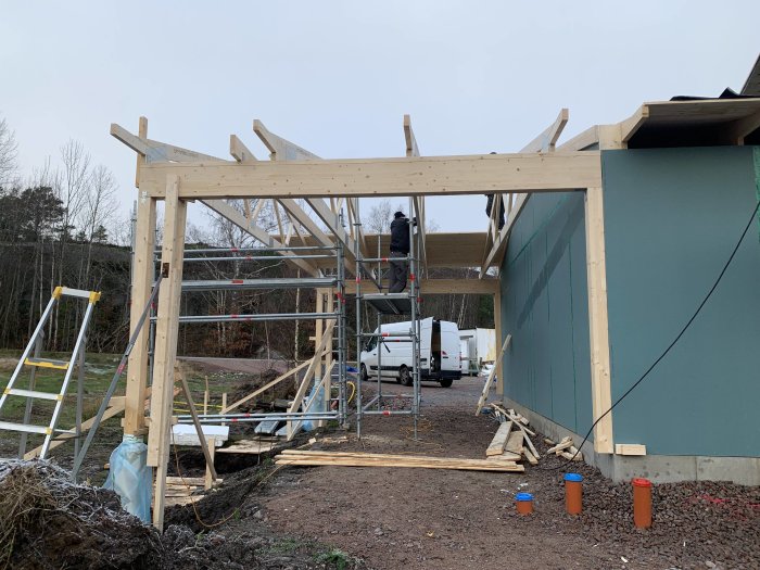 Konstruktion av trästomme för carport med arbetare på ställning och byggmaterial runtomkring.