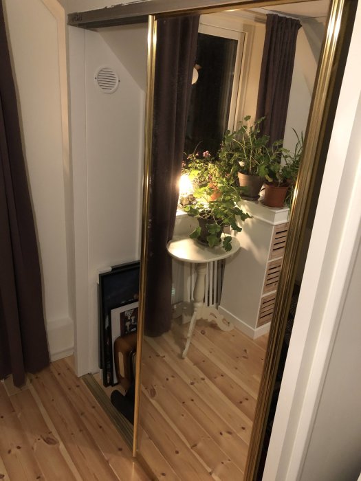 Ett hörn av ett rum med en spegel och en vit rund bord, både återspeglande och omgiven av krukväxter, bredvid en vit dörr.