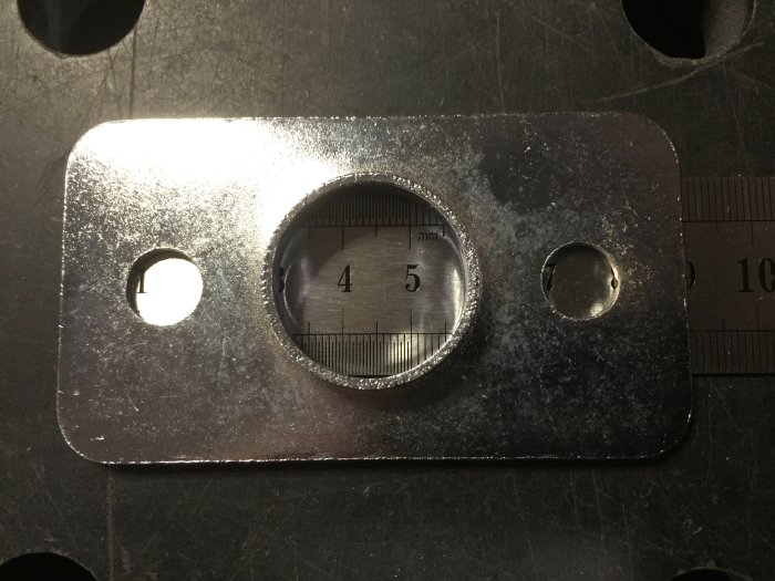 Metallplatta med ovalt hål och skala, sannolikt verktyg, bredvid skjutmått på mörk yta.