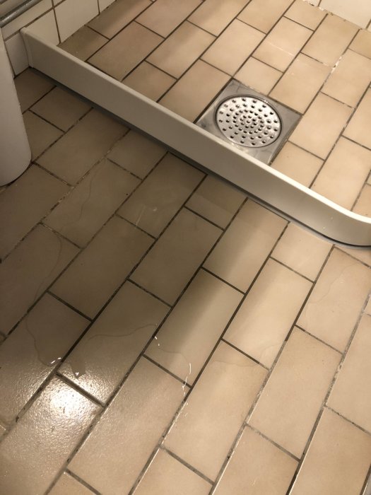 Duschkabin med felaktigt fall på golvet som leder till vattenansamling utanför duschområdet med slitna fogar.
