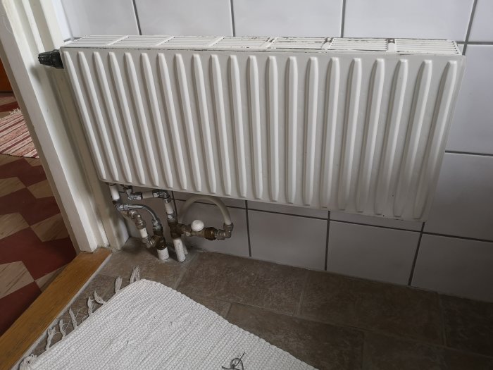 Vit radiator i ett hörn av ett badrum med synliga rör och ventiler, intill kaklad vägg.