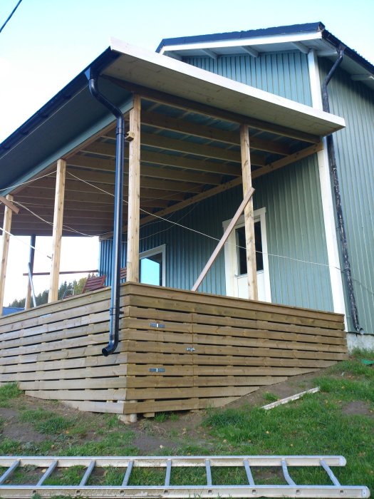 Nybyggd träveranda med tak och brun panel på ett blått hus, komplett med stuprör och fundament.