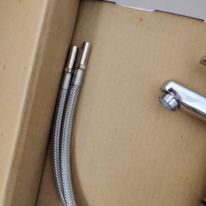 En ny blandare utan mutter och dess anslutningsslangar ligger på ett kartongunderlag.