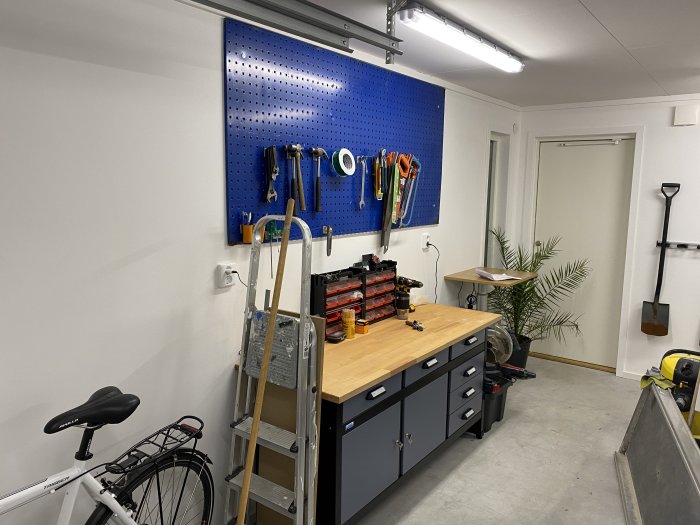 Garage med blå verktygstavla på vägg, arbetsbord, cykel, stege och element synligt i nedre kanten.