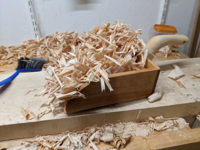 Handtillverkad träskyffel full med träspån på en arbetsbänk bredvid blå kvast.