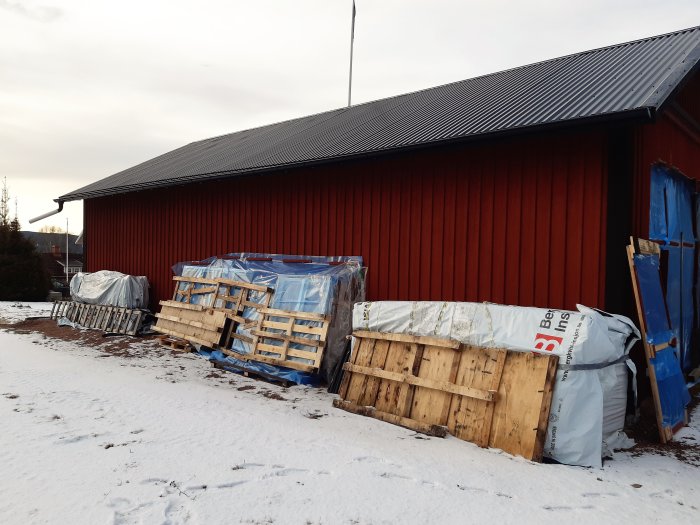 Byggmaterial och isoleringspaket framför röd verkstad under vinter.