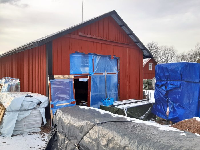 Röd verkstadsbyggnad under ombyggnad med blå presenningar och isoleringsmaterial.