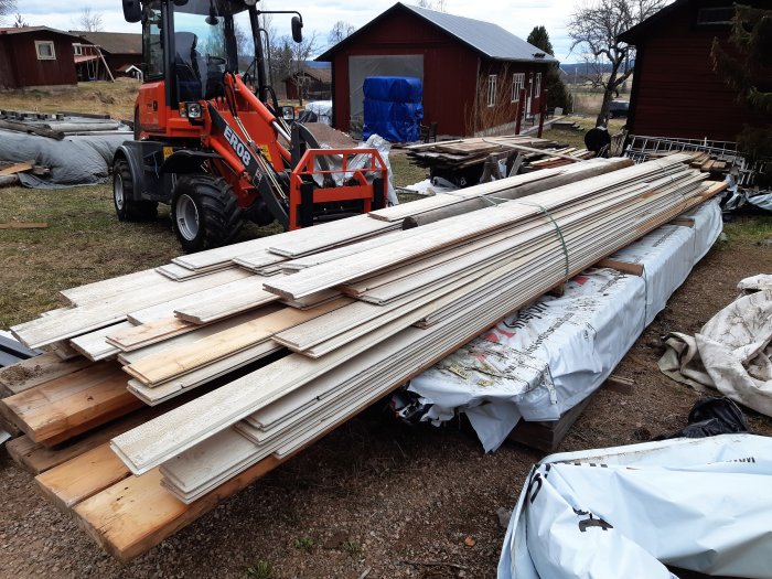 Traktor som lastar plankor och byggmaterial på en gård med byggnader i bakgrunden.