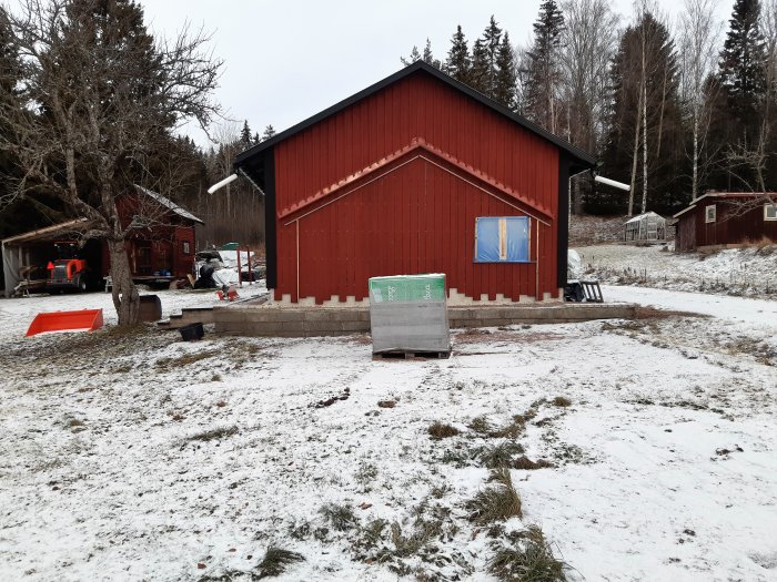 Röd verkstad med snötäckt mark och byggmaterial framför i ett vinterlandskap.