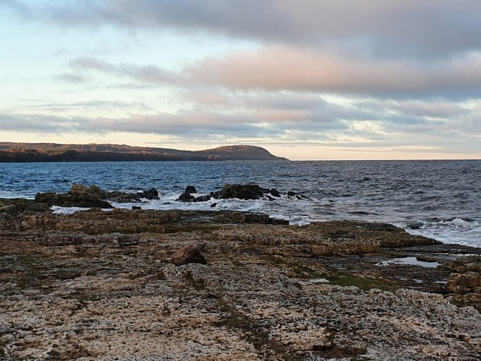 Klippig kustlinje med vågor som slår mot stranden under en molnig himmel vid skymning.