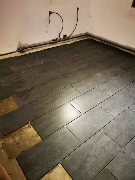 Elledningar och rör på en vägg ovanför ett delvis kaklat golv med synliga hål och gamla eluttaget.