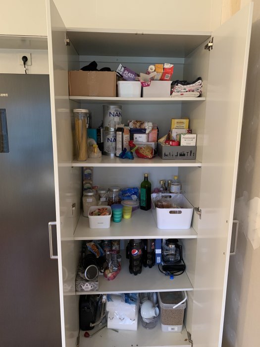 Öppet högskåp fyllt med oorganiserade matvaror och köksredskap i behov av trådbackar.