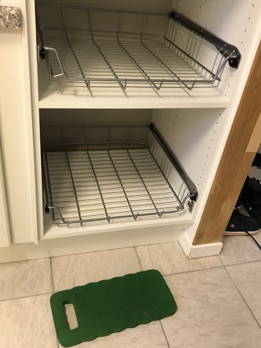 Öppet köksskåp med två utdragbara trådbackar Utrusta installerade, grönt skärbräde på golvet framför.