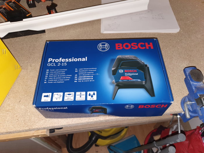 Förpackning av Bosch Professional GCL 2-15 krysslaser på ett arbetsbord med ritningar och verktyg.