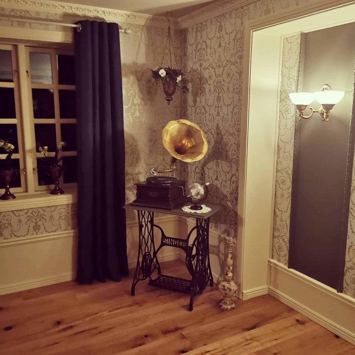 Hörn av ett tv-rum med ett gramofon på ett bord, mörka gardiner och tapet med mönster, snyggt upplyst av en vägglampa.