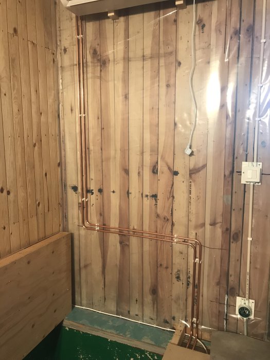 Installation av en fläktkonvektor med kopparledningar på en trävägg i ett rum under renovering.