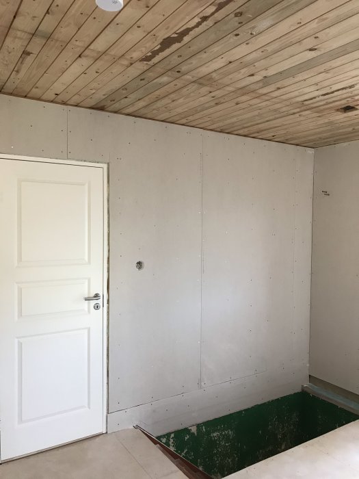 Rum under renovering med nyligen monterade gipsskivor på väggen, träpanel i taket och vit dörr.