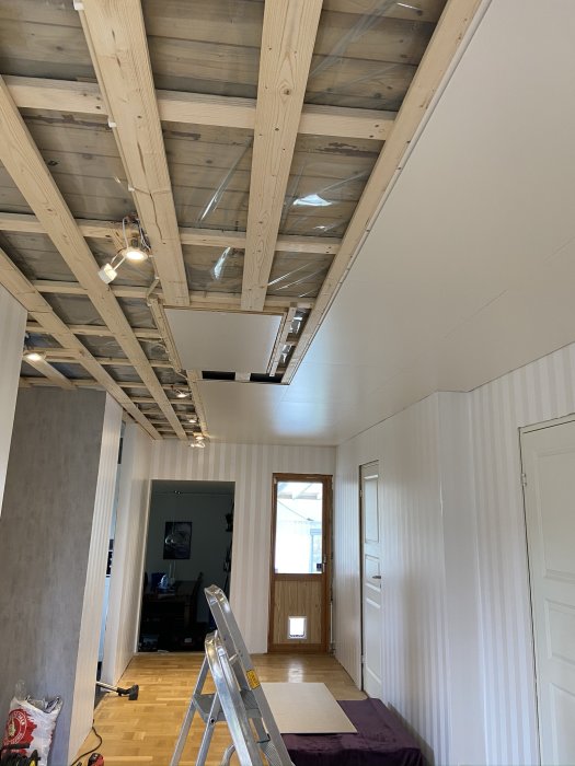 Inomhus byggarbetsplats med oskyddade takbjälkar och en öppen plats för innertaket, stege i förgrunden.