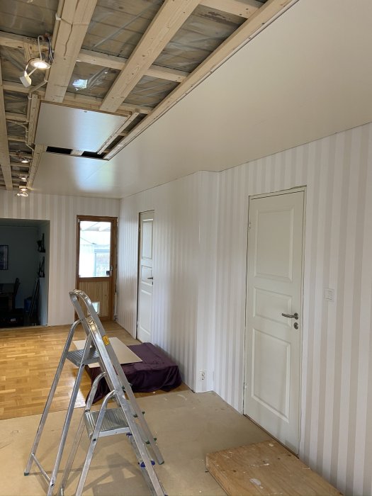 Rum under renovering med öppet innertak i väntan på installation av spotlights, stege framför vägg.