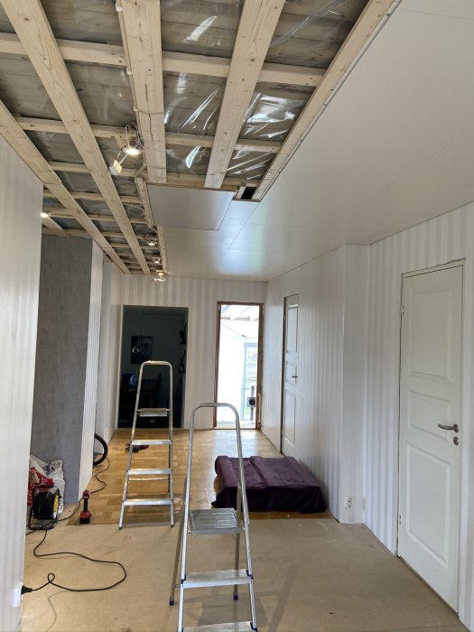 Delvis installerat innertak med öppna träbjälkar och en monterad spotlight, stege och byggmaterial på golvet.